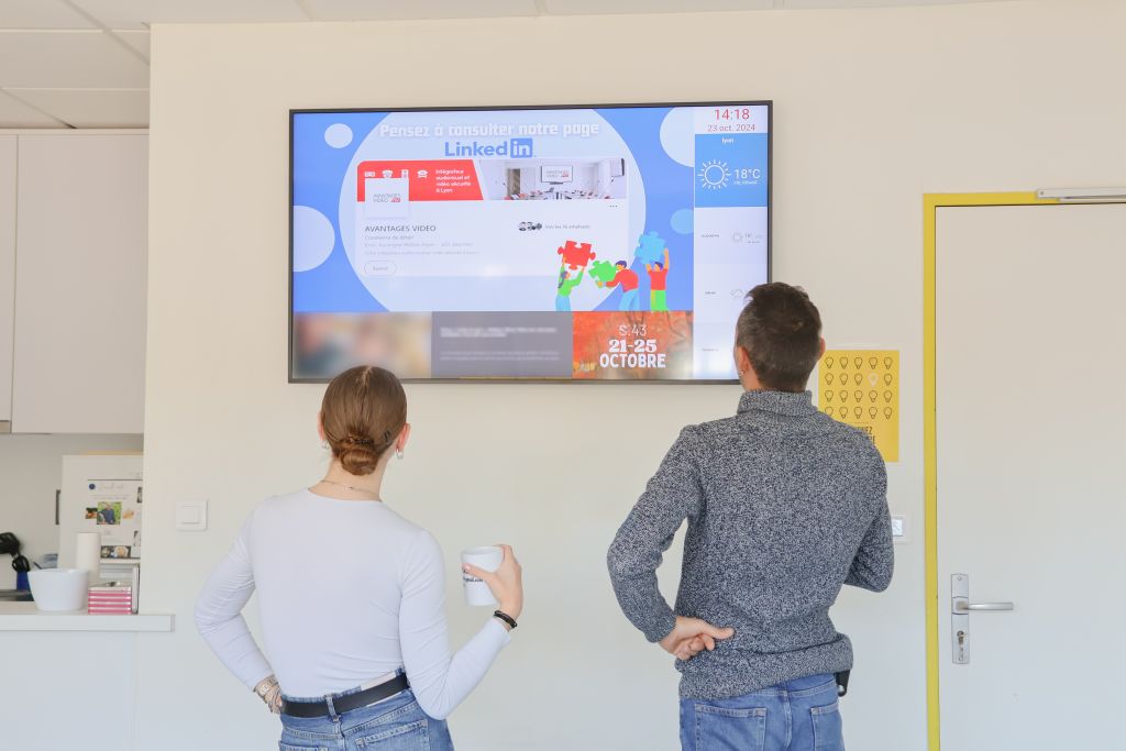 Écran avec affichage dynamique dans une salle de pause où deux employés prennent le café en consultant les informations du jour sur l'écran. Ces informations défilent selon les actualités.
